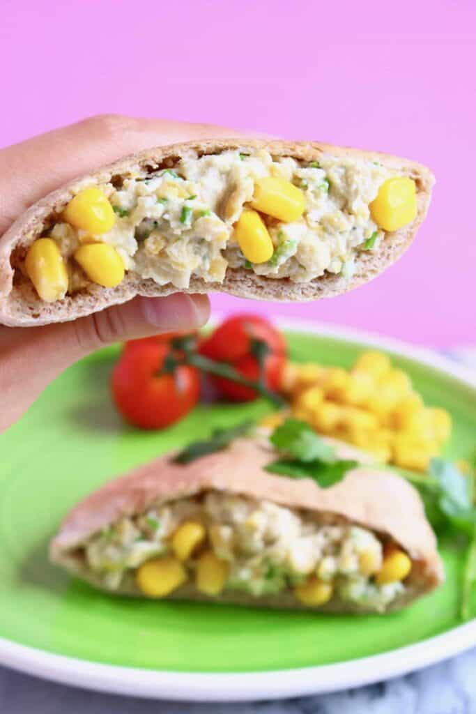 Half a pitta bread filled with creamy chickpeas and yellow sweetcorn being held up with a hand against a pink background