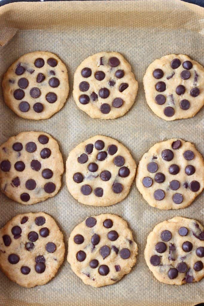 Nine raw chocolate chip cookies on a baking tray lined with brown baking paper