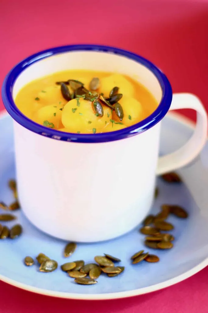 Photo of orange soup with gnocchi topped with green pumpkin seeds in a white mug with a dark blue rim on a light blue plate scattered with pumpkin seeds against a bright pink background