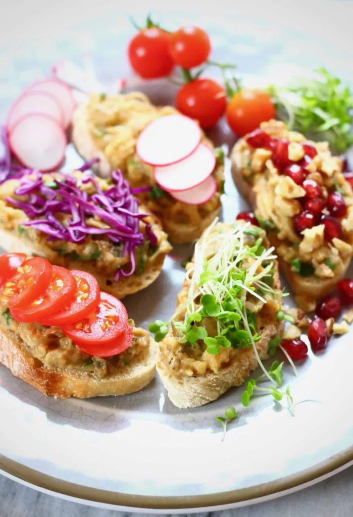 Five pieces of bruschetta topped with shredded eggplant with pomegranate seeds, shredded purple cabbage, cress, sliced radish, sliced tomatoes on a grey plate