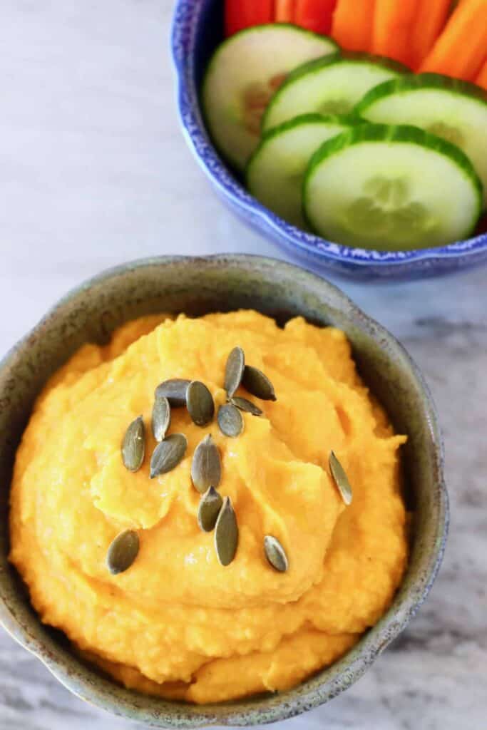 Photo of a grey bowl of orange pumpkin hummus topped with green pumpkin seeds and a blue bowl of chopped vegetables on a marble surface