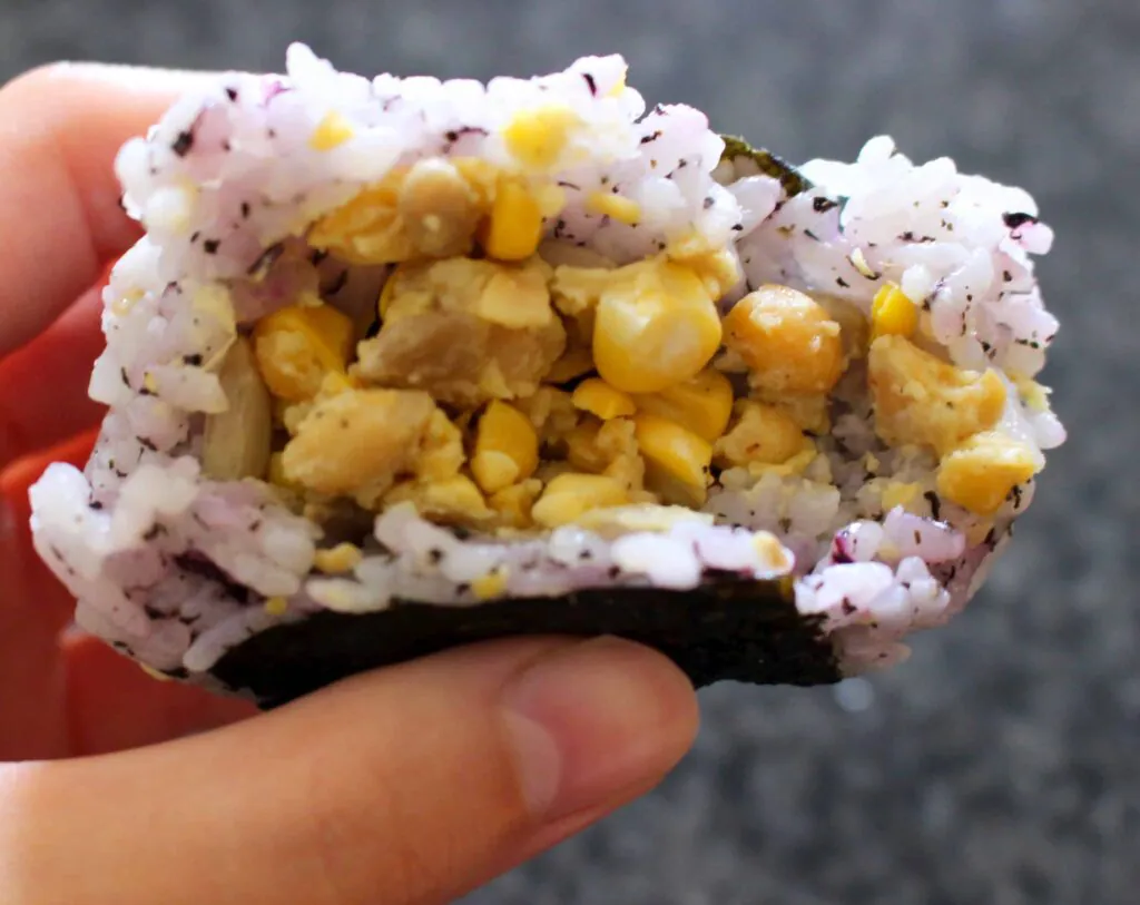 An onigiri filled with mashed chickpeas and sweetcorn being held up with a hand against a dark grey background