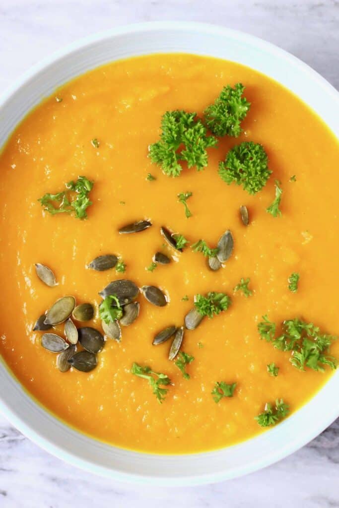 Photo of smooth orange pumpkin soup topped with green pumpkin seeds and curly parsley in a light blue bowl against a marble background