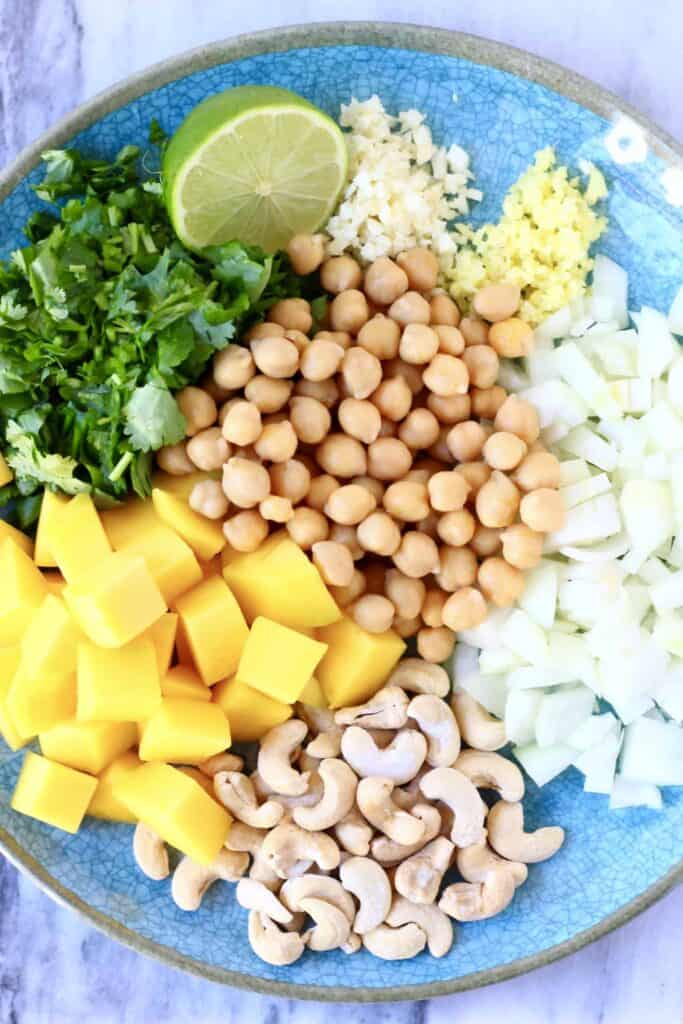 Diced mango, cashew nuts, chickpeas, chopped coriander, half a lime, minced garlic, minced garlic and diced onion on a blue plate against a marble background