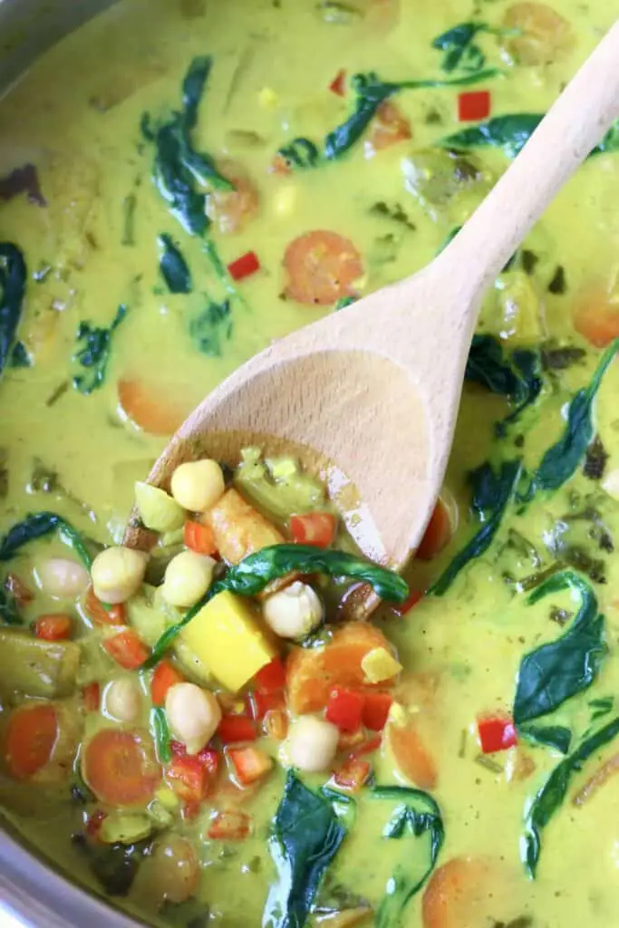 Yellow curry made using diced mango, chickpeas, carrots and spinach in a silver pan being stirred with a wooden spoon