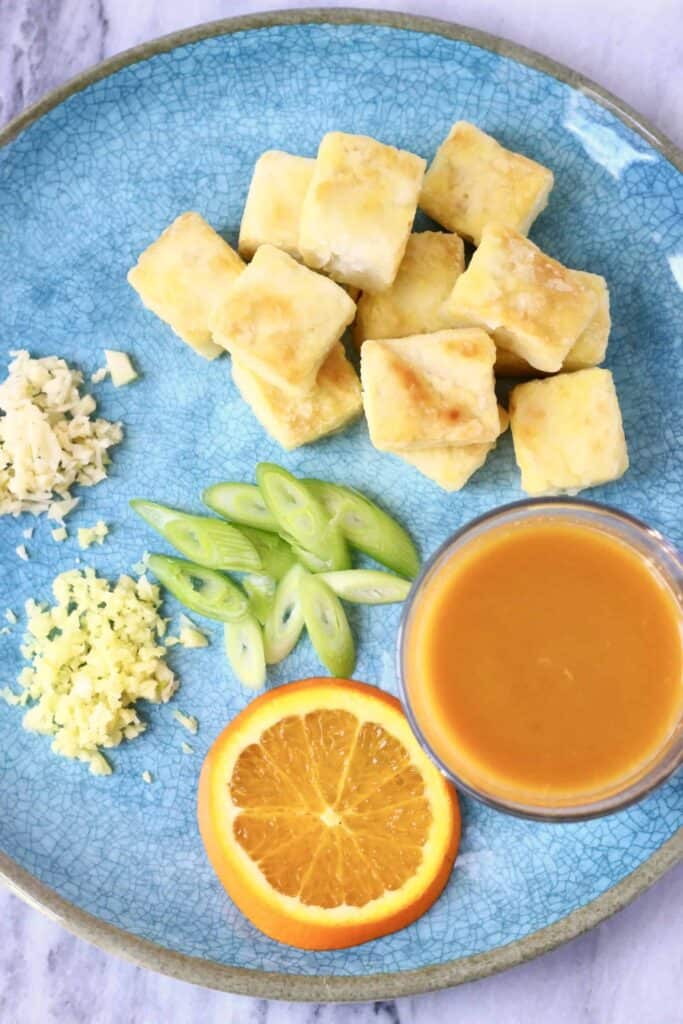 Cubes of brown fried tofu with minced garlic, ginger, sliced spring onions, a slice of orange and a glass of orange juice on a blue plate against a marble background