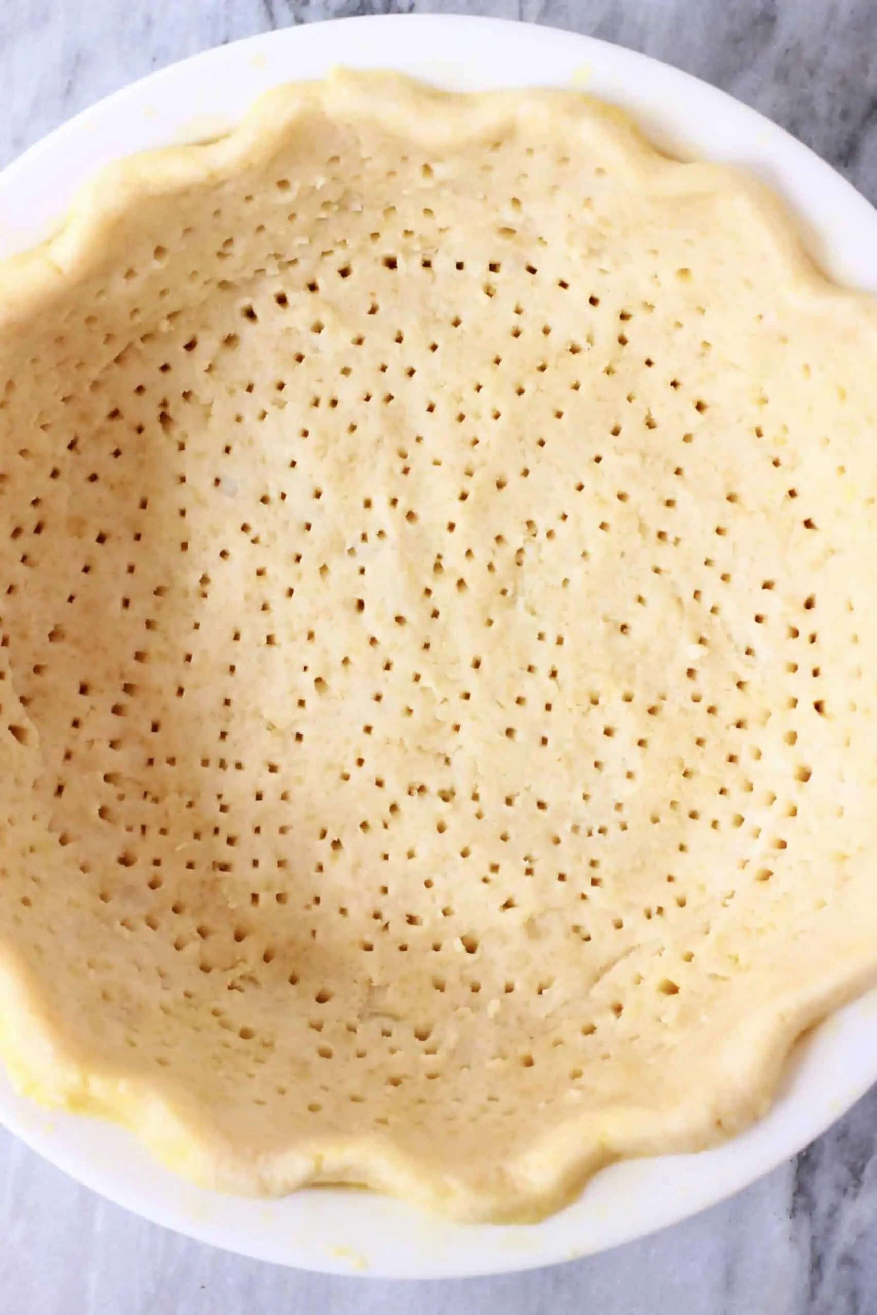 Photo of a pie crust with lots of holes poked in the surface in a white pie dish against a marble background