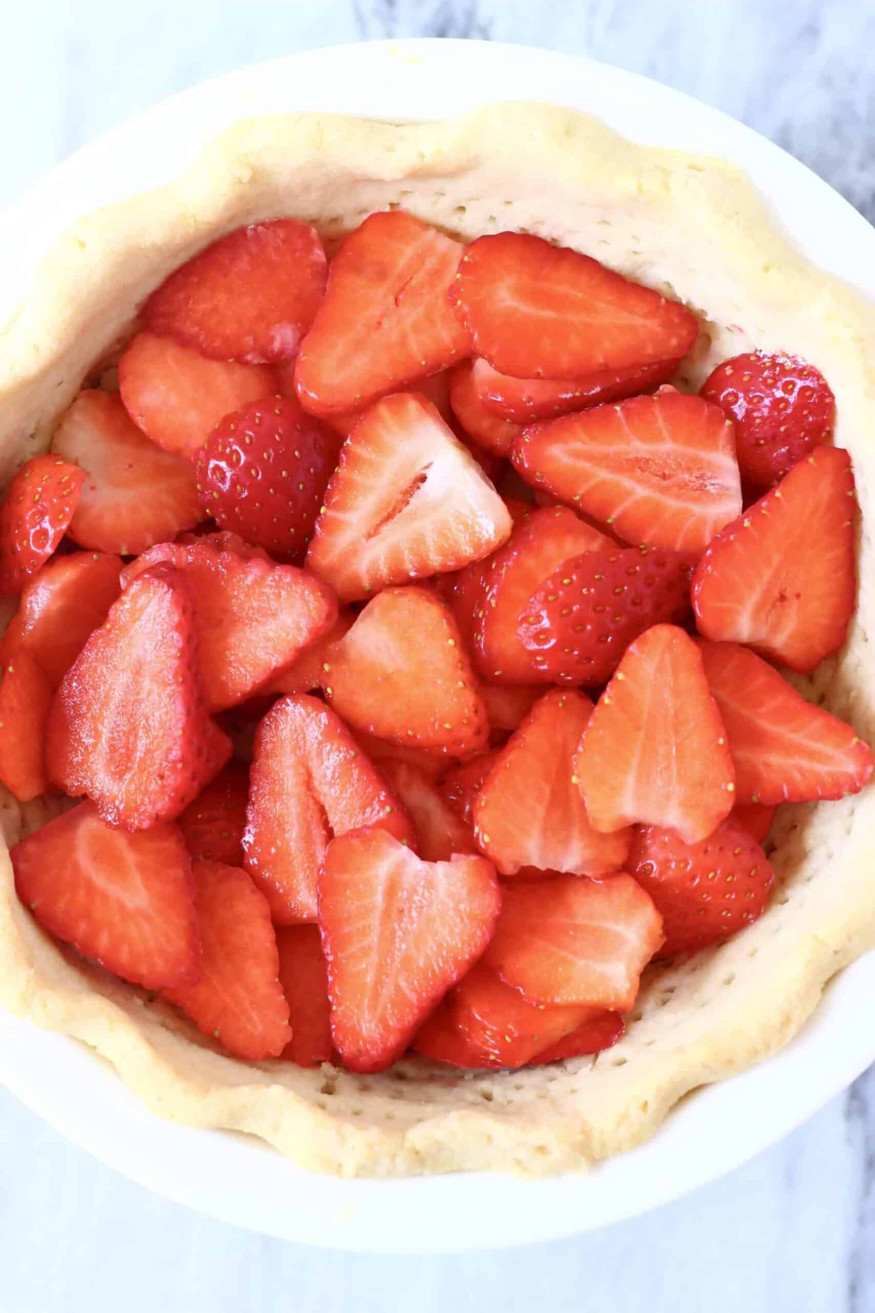 This Gluten-Free Vegan Strawberry Pie is crispy and flaky, fresh and fruity, and sweet and juicy! Refined sugar free and naturally coloured. A pie crust filled with sliced strawberries and agar fresh strawberry jelly. Perfect for summer entertaining - BBQs, potlucks, picnics, baby showers etc. #vegan #glutenfree #dairyfree #plantbased #pie #dessert #strawberry #summer Photo of a pie crust filled with sliced strawberries in a white pie dish against a marble background