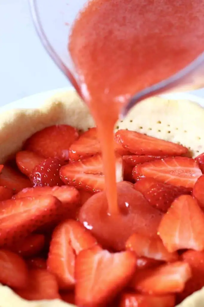 Photo of a pie crust filled with sliced strawberries with a jug pouring strawberry sauce into it 
