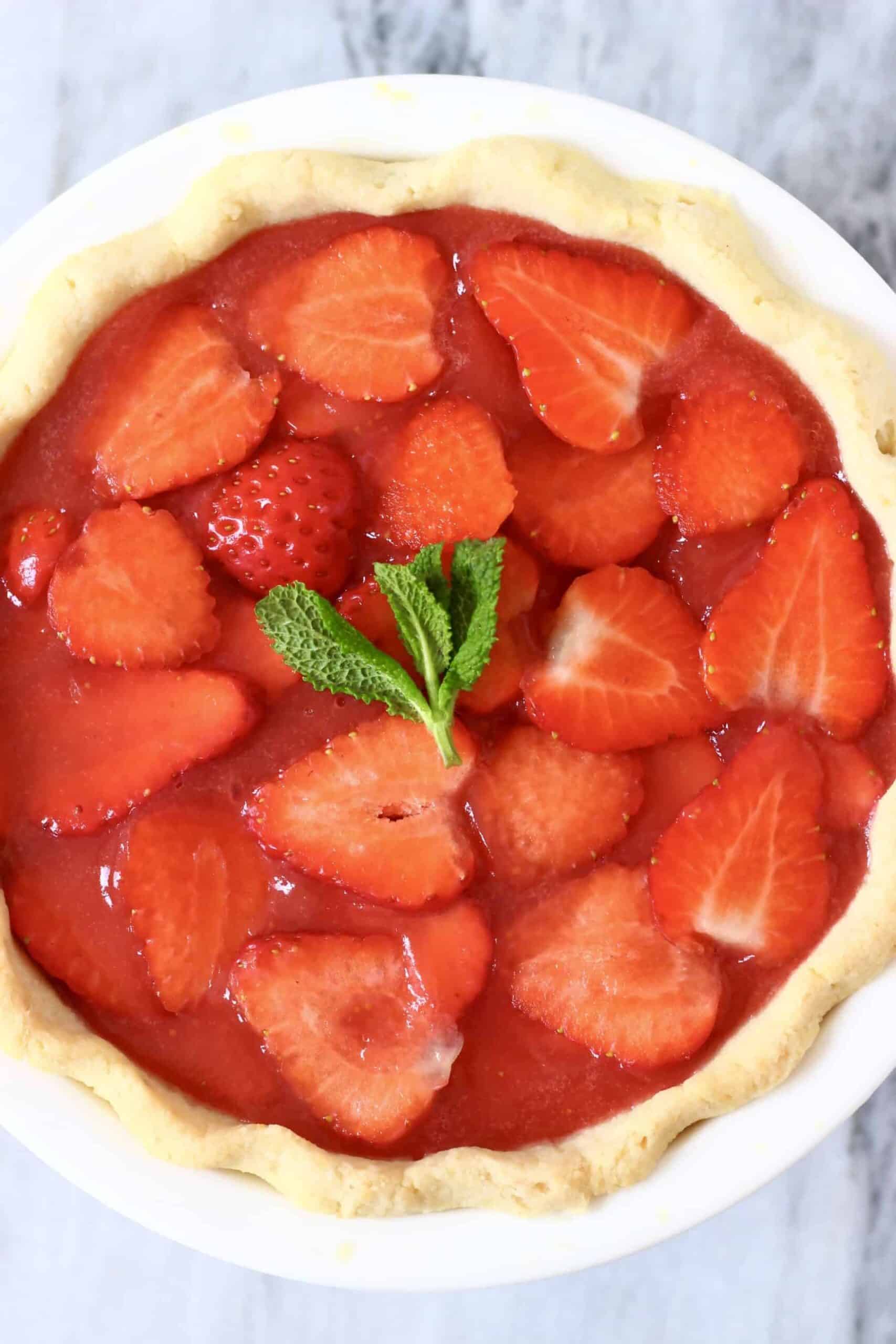 Photo of a strawberry pie topped with a sprig of mint in a white pie dish against a marble background