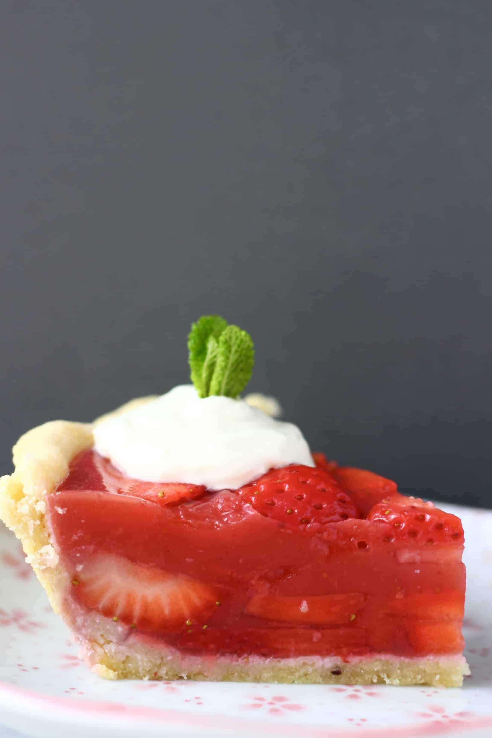 This Gluten-Free Vegan Strawberry Pie is crispy and flaky, fresh and fruity, and sweet and juicy! Refined sugar free and naturally coloured. A pie crust filled with sliced strawberries and agar fresh strawberry jelly. Perfect for summer entertaining - BBQs, potlucks, picnics, baby showers etc. #vegan #glutenfree #dairyfree #plantbased #pie #dessert #strawberry #summer Photo of a slice of strawberry pie topped with whipped cream and a sprig of mint on a pink plate against a grey background