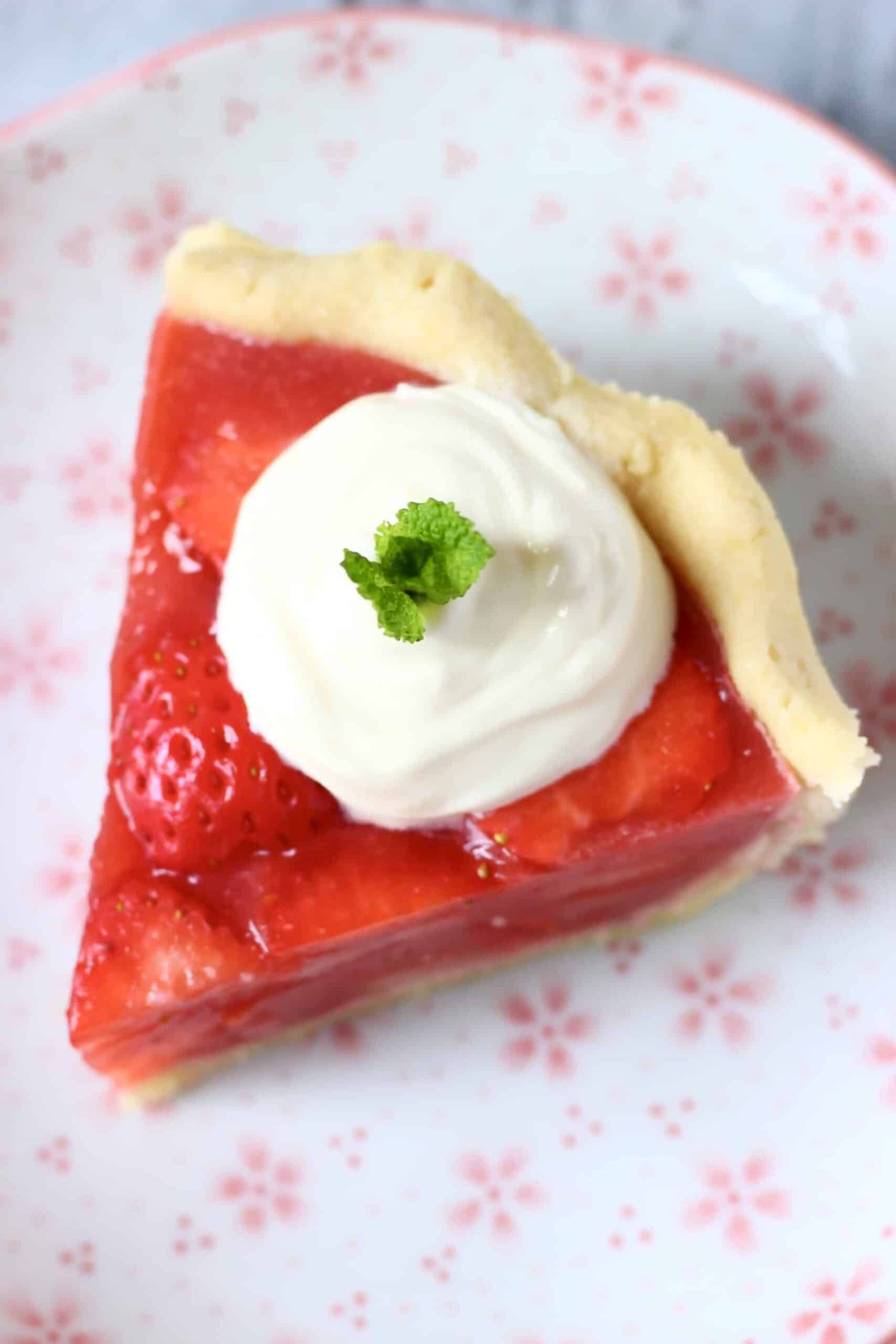 This Gluten-Free Vegan Strawberry Pie is crispy and flaky, fresh and fruity, and sweet and juicy! Refined sugar free and naturally coloured. A pie crust filled with sliced strawberries and agar fresh strawberry jelly. Perfect for summer entertaining - BBQs, potlucks, picnics, baby showers etc. #vegan #glutenfree #dairyfree #plantbased #pie #dessert #strawberry #summer Photo of a slice of strawberry pie topped with cream and a sprig of mint against a white plate with pink flowers