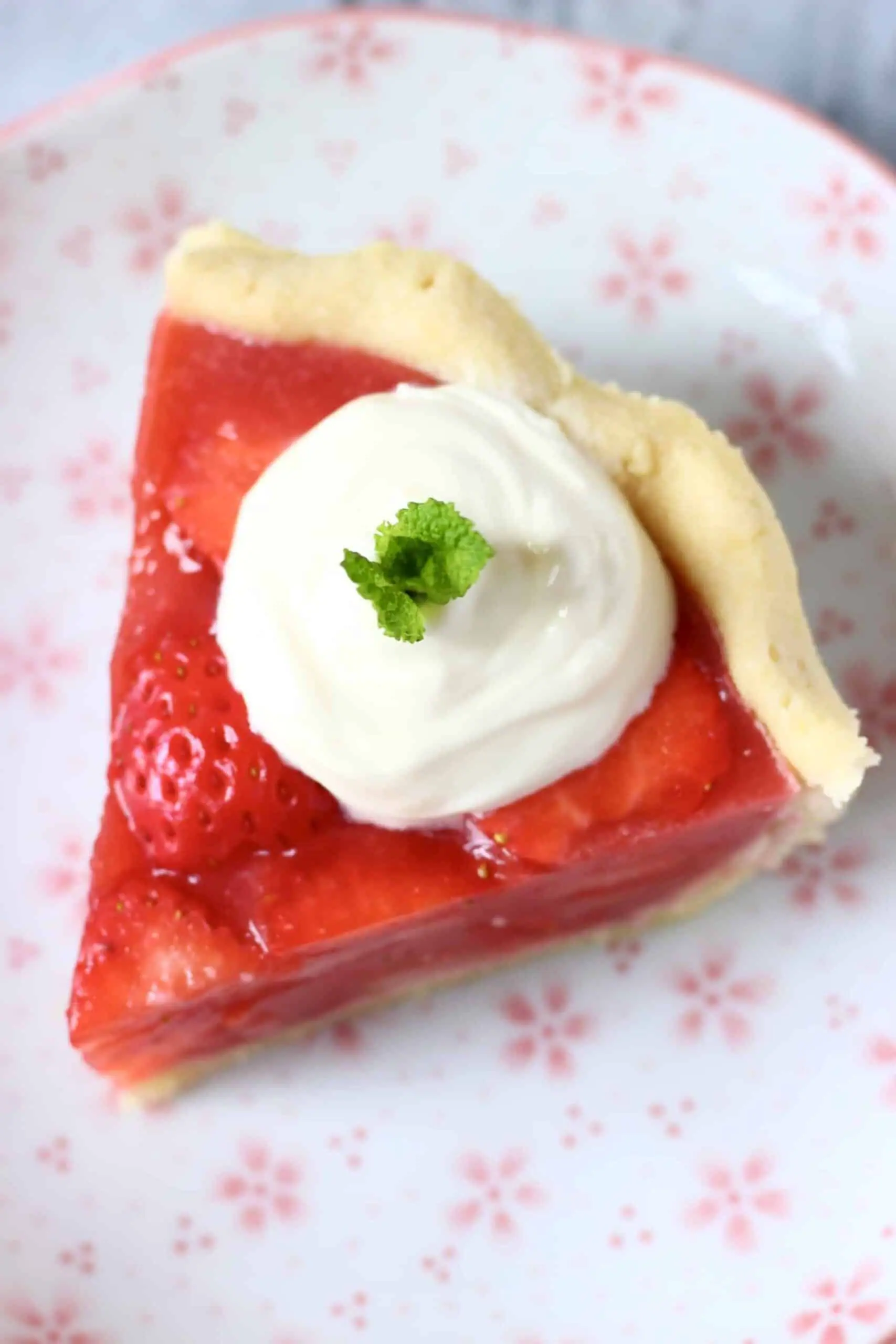 Photo of a slice of strawberry pie topped with cream and a sprig of mint against a white plate with pink flowers