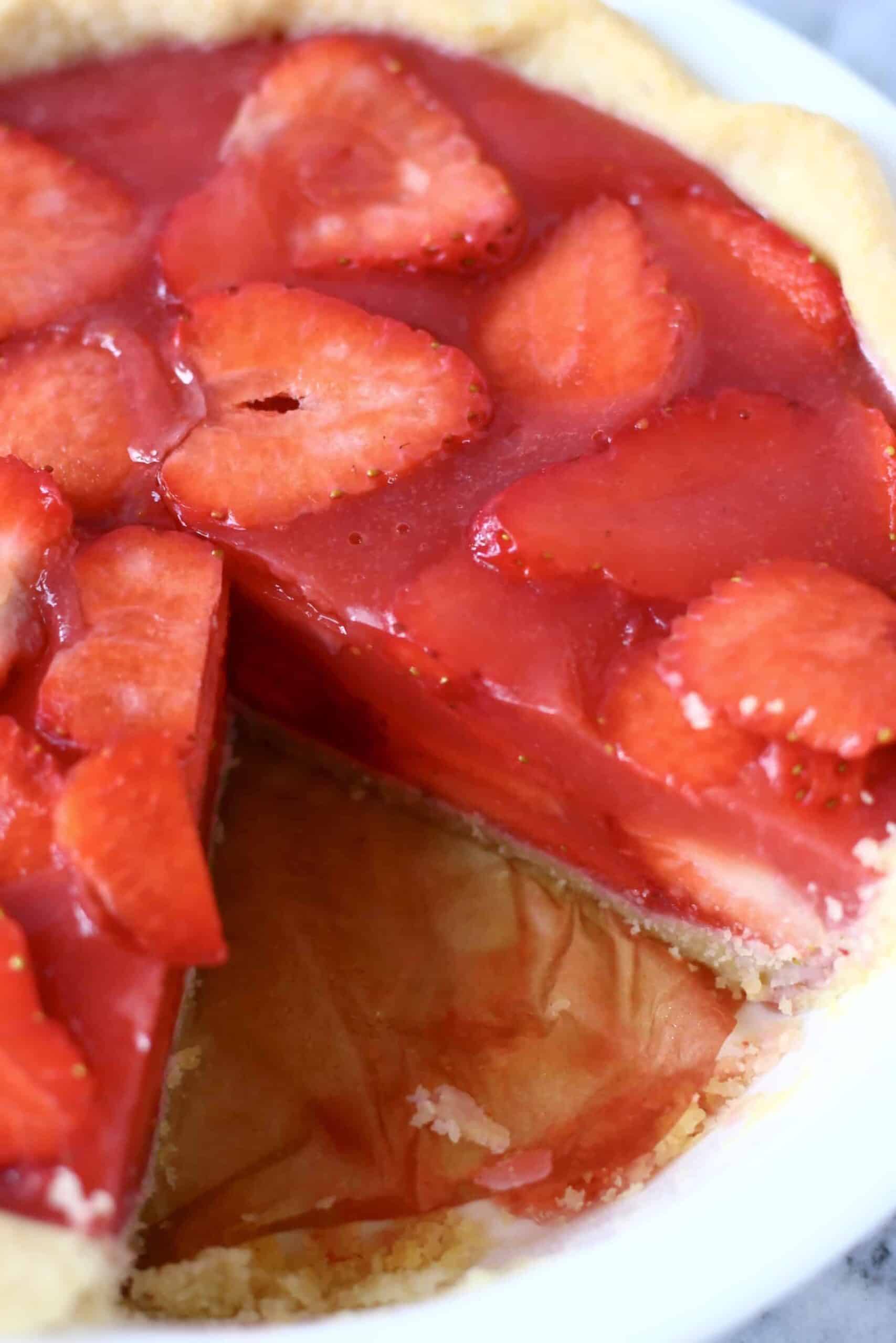 This Gluten-Free Vegan Strawberry Pie is crispy and flaky, fresh and fruity, and sweet and juicy! Refined sugar free and naturally coloured. A pie crust filled with sliced strawberries and agar fresh strawberry jelly. Perfect for summer entertaining - BBQs, potlucks, picnics, baby showers etc. #vegan #glutenfree #dairyfree #plantbased #pie #dessert #strawberry #summer Photo of a strawberry pie with a slice cut out of it in a white pie dish