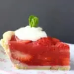 Photo of a slice of strawberry pie topped with whipped cream and a sprig of mint on a pink plate against a grey background