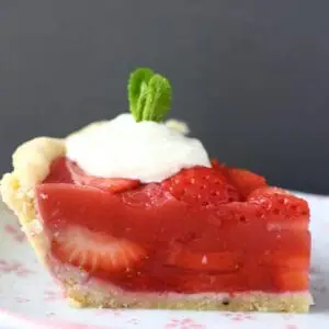 Photo of a slice of strawberry pie topped with whipped cream and a sprig of mint on a pink plate against a grey background