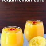 Two glasses filled with bright yellow lemon curd topped with freeze-dried raspberries on a marble surface with half a squeezed lemon against a dark brown background