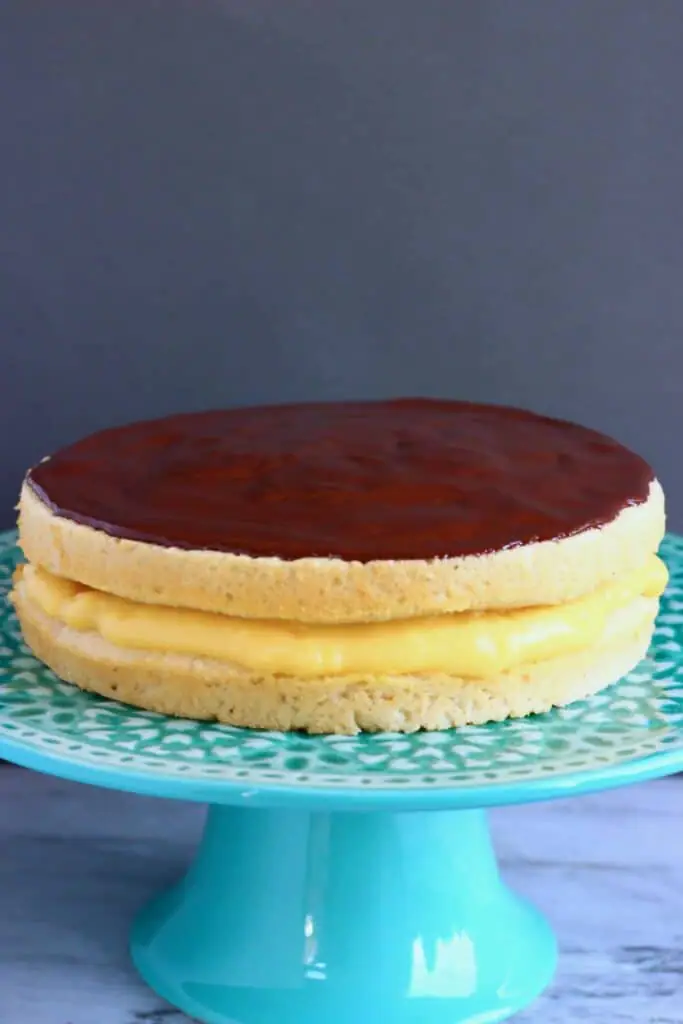 A golden brown sponge cake filled with yellow custard topped with chocolate ganache on a green cake stand against a grey background