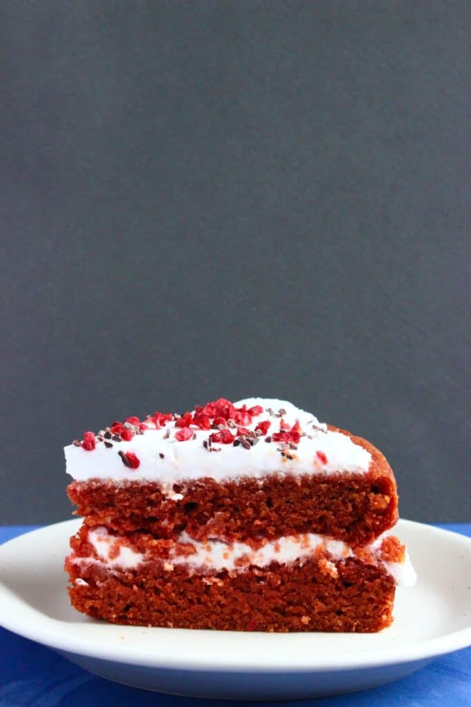 Photo of a slice of red sponge cake with creamy white frosting on a small white plate against a grey background