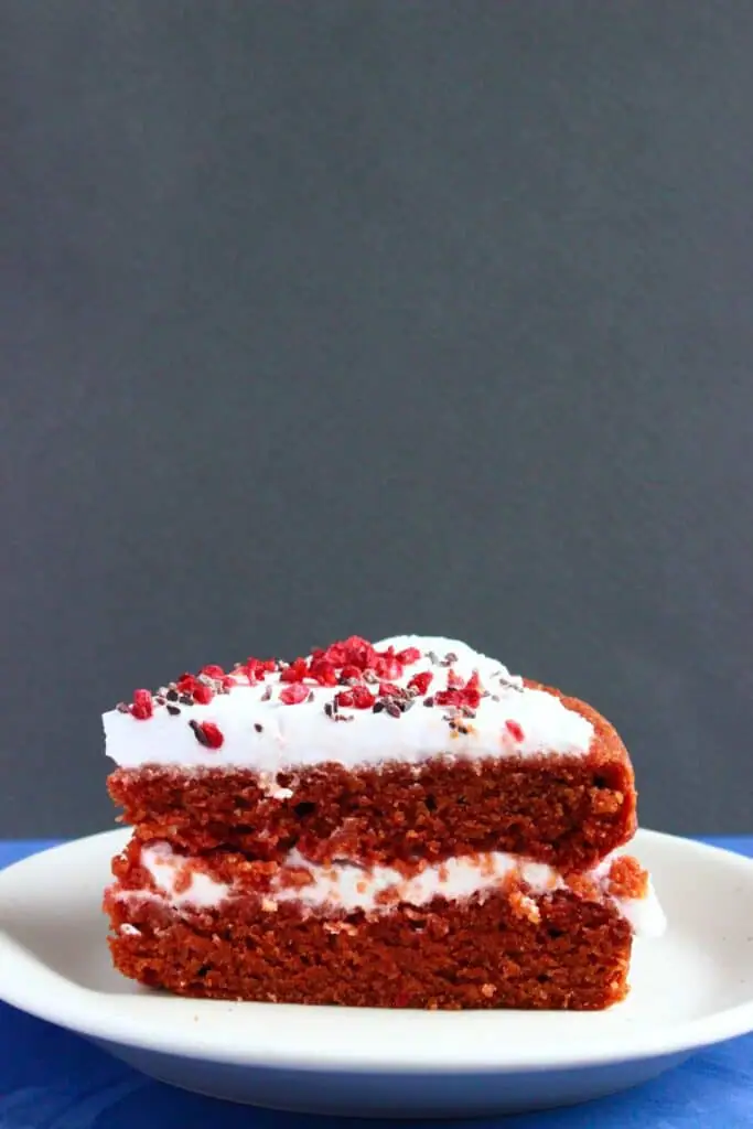 Photo of a slice of red sponge cake with creamy white frosting on a small white plate against a grey background