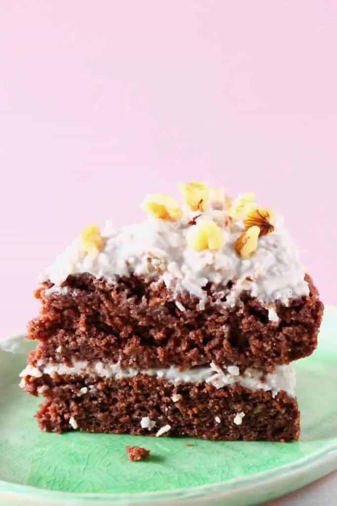 A slice of chocolate sponge topped with creamy white frosting and walnuts on a green plate against a pink background