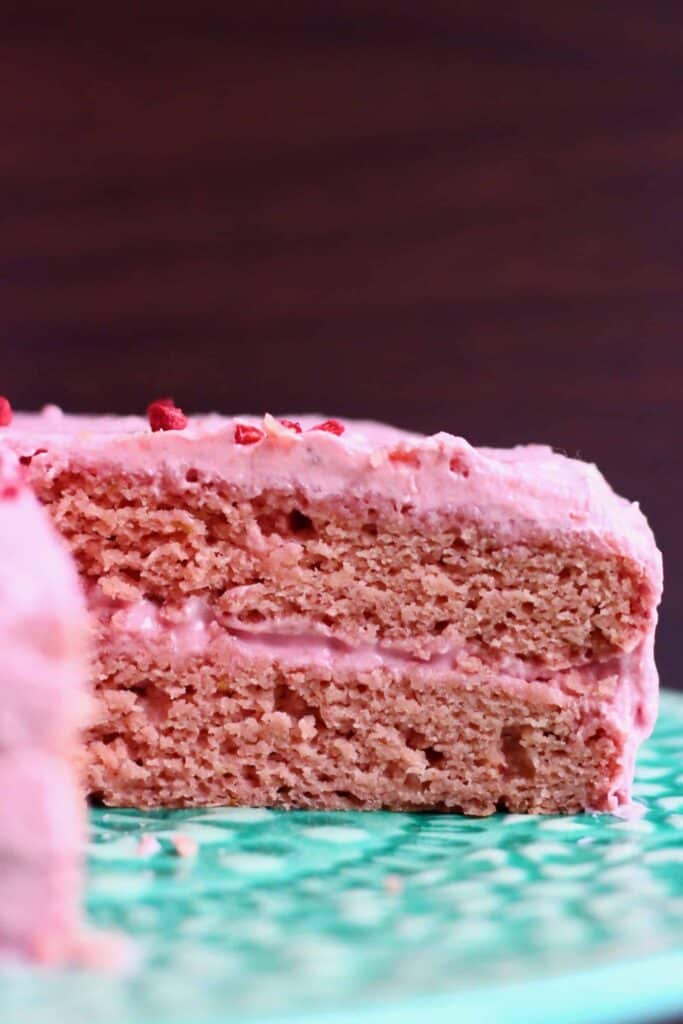 Photo of a sliced pink sponge cake with pink frosting on a green cake stand against a dark brown background