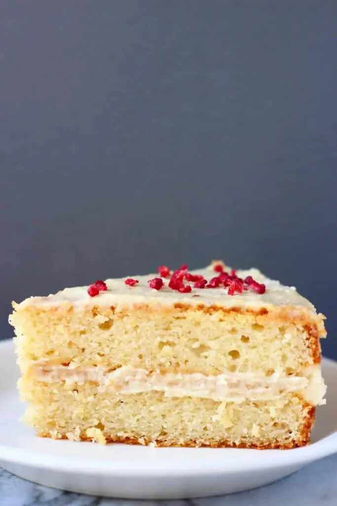 Photo of a slice of yellow sponge cake sandwiched with white buttercream topped with freeze-dried raspberries on a white plate against a grey background
