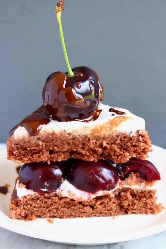 Photo of a slice of chocolate cake frosted with white creamy frosting topped with a black cherry and chocolate sauce on a white plate against a grey background