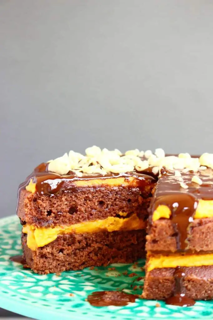 Photo of chocolate cake sandwiched with yellow peanut butter frosting, drizzled with chocolate ganache and sprinkled with chopped peanuts on a green cake stand against a grey background