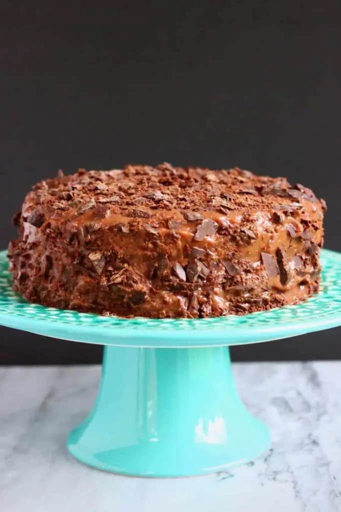 Photo of a chocolate cake covered in bits of chopped up chocolate on a blue cake stand against a grey background