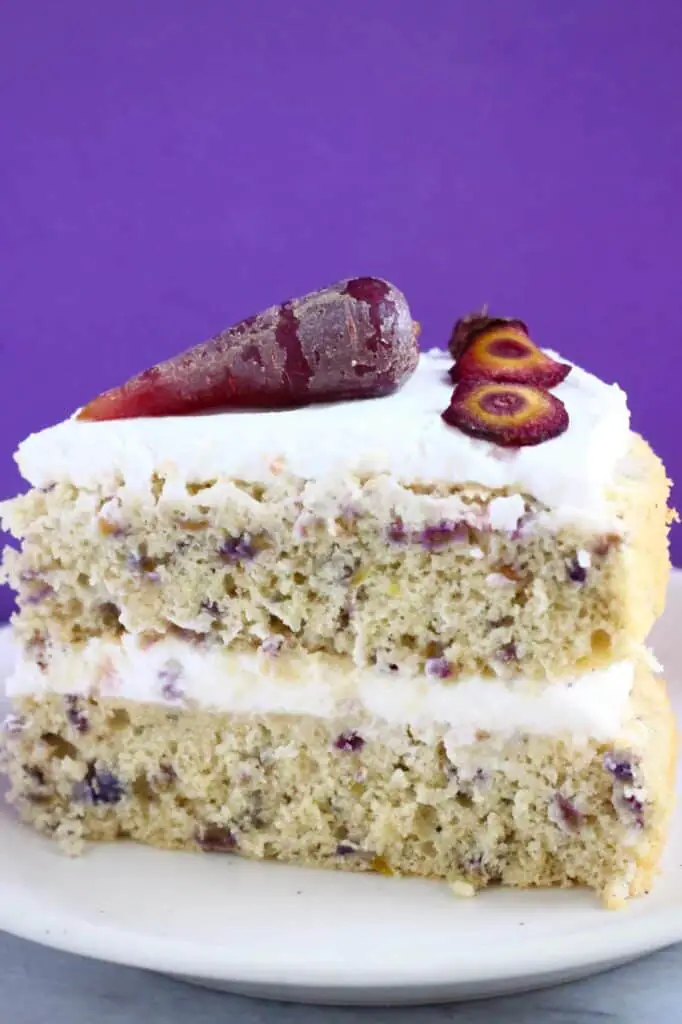 A slice of carrot cake made with purple carrots topped with a purple carrot, on a small white plate against a purple background