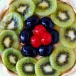 Photo of a fruit tart topped with sliced kiwi, grapes and cherries in a white pie dish against a marble background