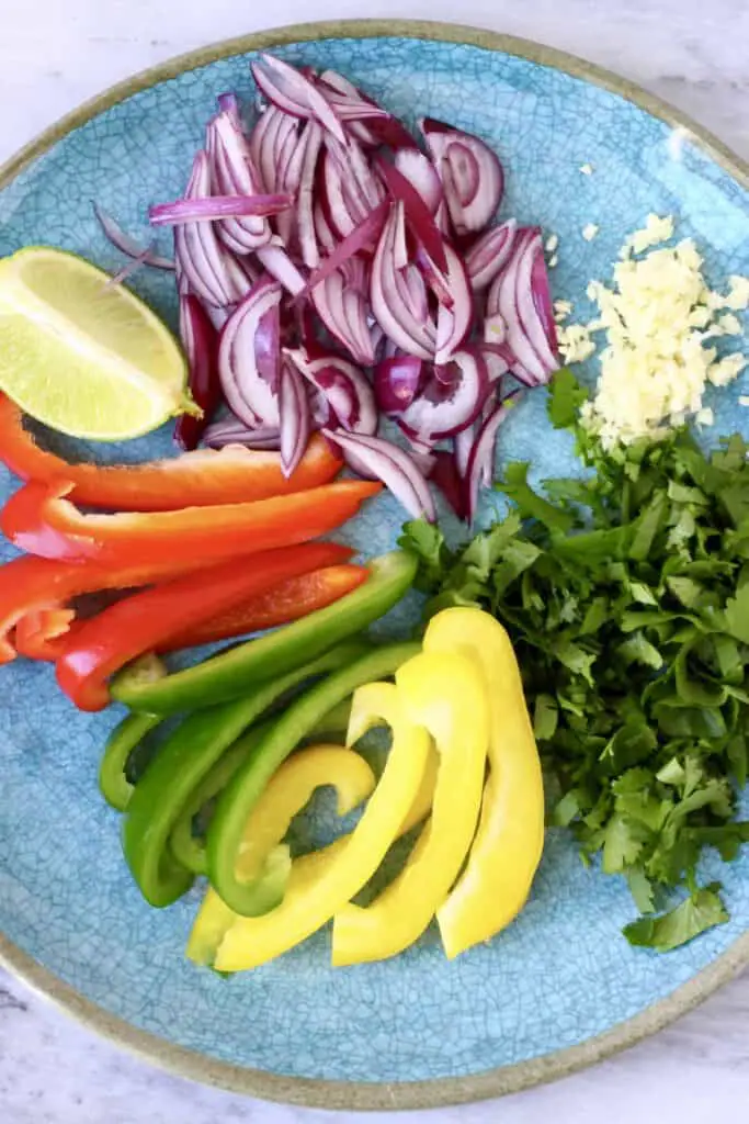 Sliced peppers, a lime wedge, chopped coriander, minced garlic and sliced red onion on a blue plate against a marble background