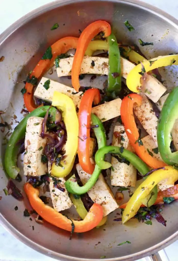 Cubes of tofu and sliced peppers in a silver frying pan with spices and herbs against a marble background