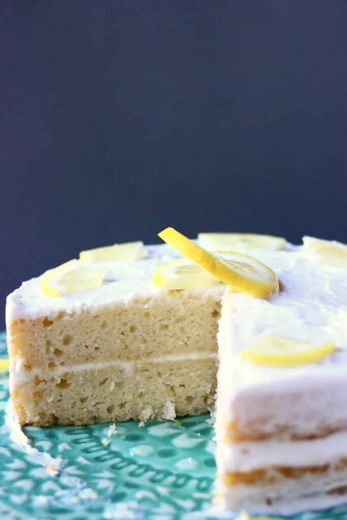 Photo of a sliced sponge cake with white frosting and a lemon slice on top on a green cake stand with a grey background