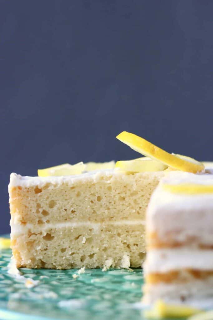 Photo of a sliced sponge cake with white frosting and a lemon slice on top on a green cake stand with a grey background