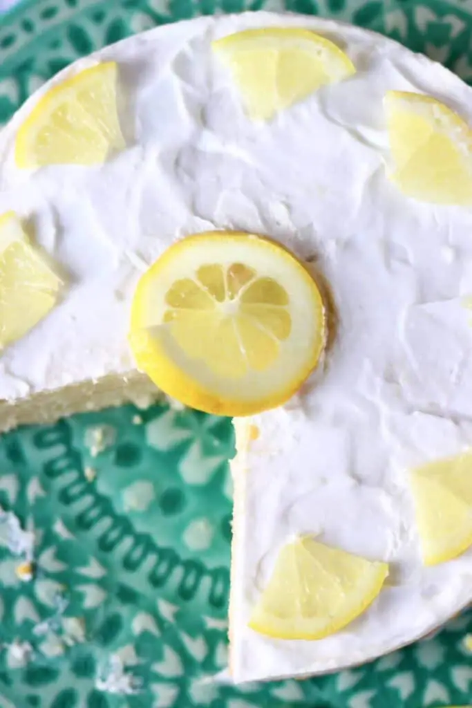 Photo of a sponge cake topped with white frosting, a lemon slice and lemon wedges on a green cake stand taken from above