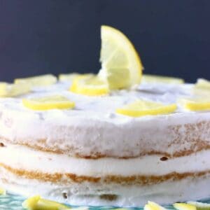 Photo of a sponge cake frosted with white creamy frosting and topped with lemon slices on a green cake stand with a grey background