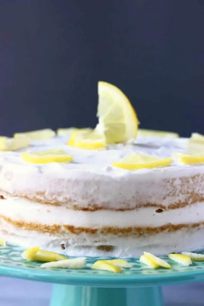 Photo of a sponge cake frosted with white creamy frosting and topped with lemon slices on a green cake stand with a grey background