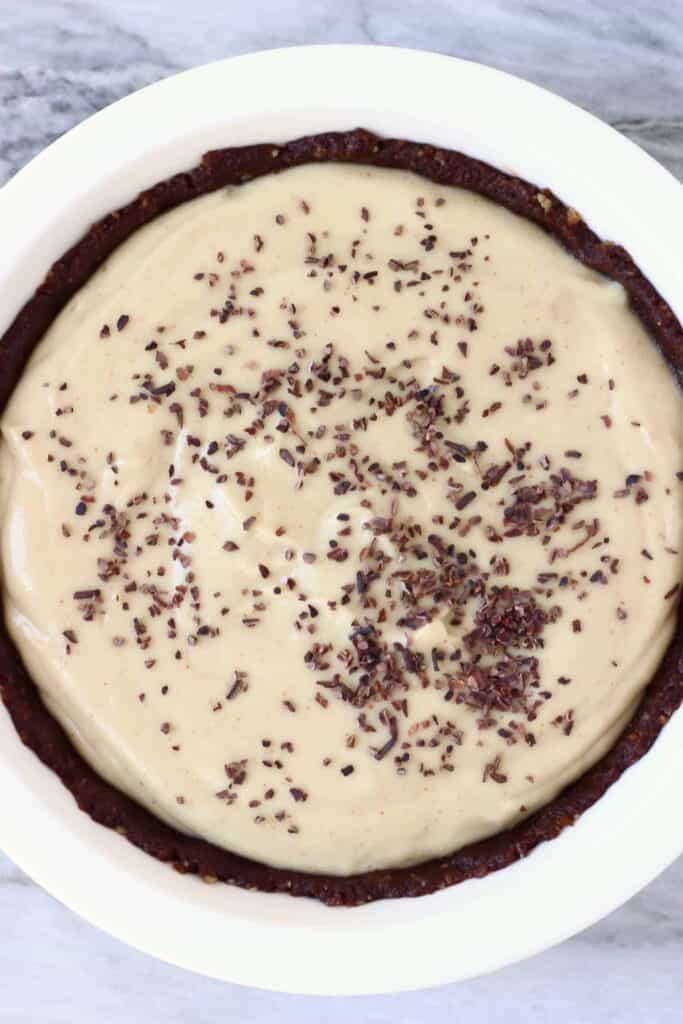 Photo of a brown chocolate pie crust filled with light brown peanut butter filling topped with cacao nibs in a white pie dish against a marble background