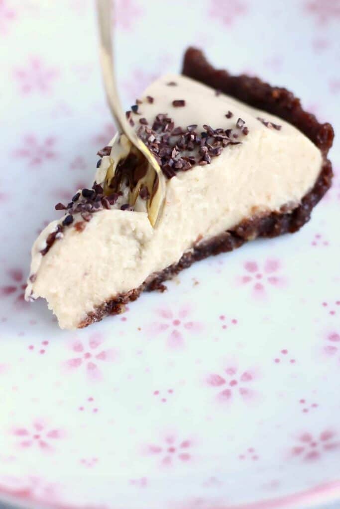 A slice of peanut butter pie with a chocolate crust and a light brown peanut butter filling sprinkled with cacao nibs with a gold fork going into the pie against a white plate with pink flowers