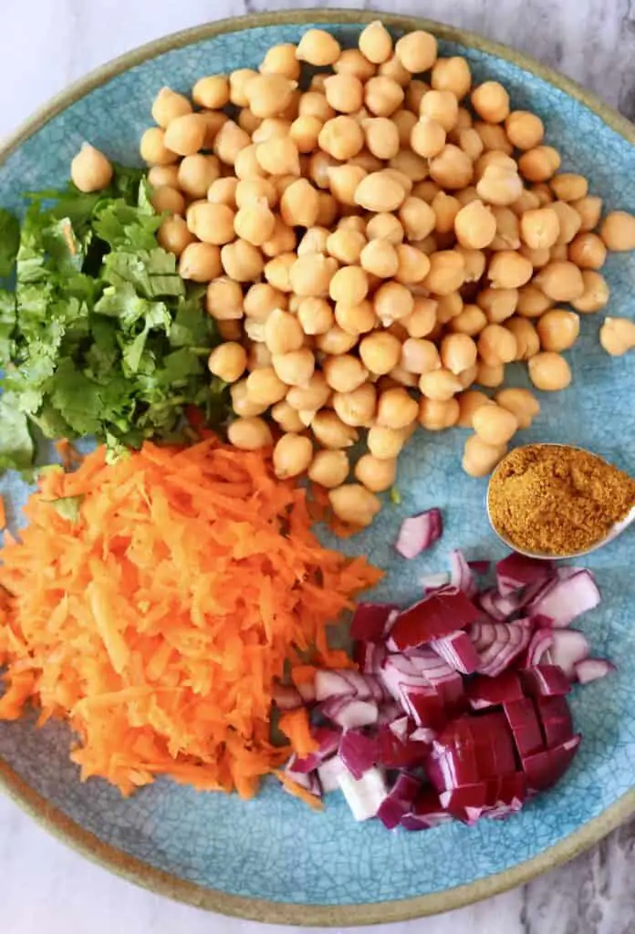 Photo of chickpeas, chopped coriander, grated carrot, diced red onion and curry powder on a blue plate against a marble background