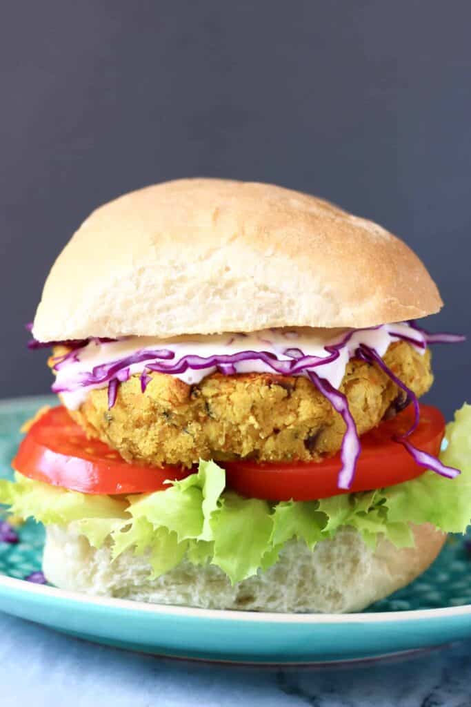 Photo of a curried chickpea burger with a bun, tomato, lettuce and purple cabbage against a grey background