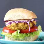 Photo of a curried chickpea burger with a bun, tomato, lettuce and purple cabbage against a grey background