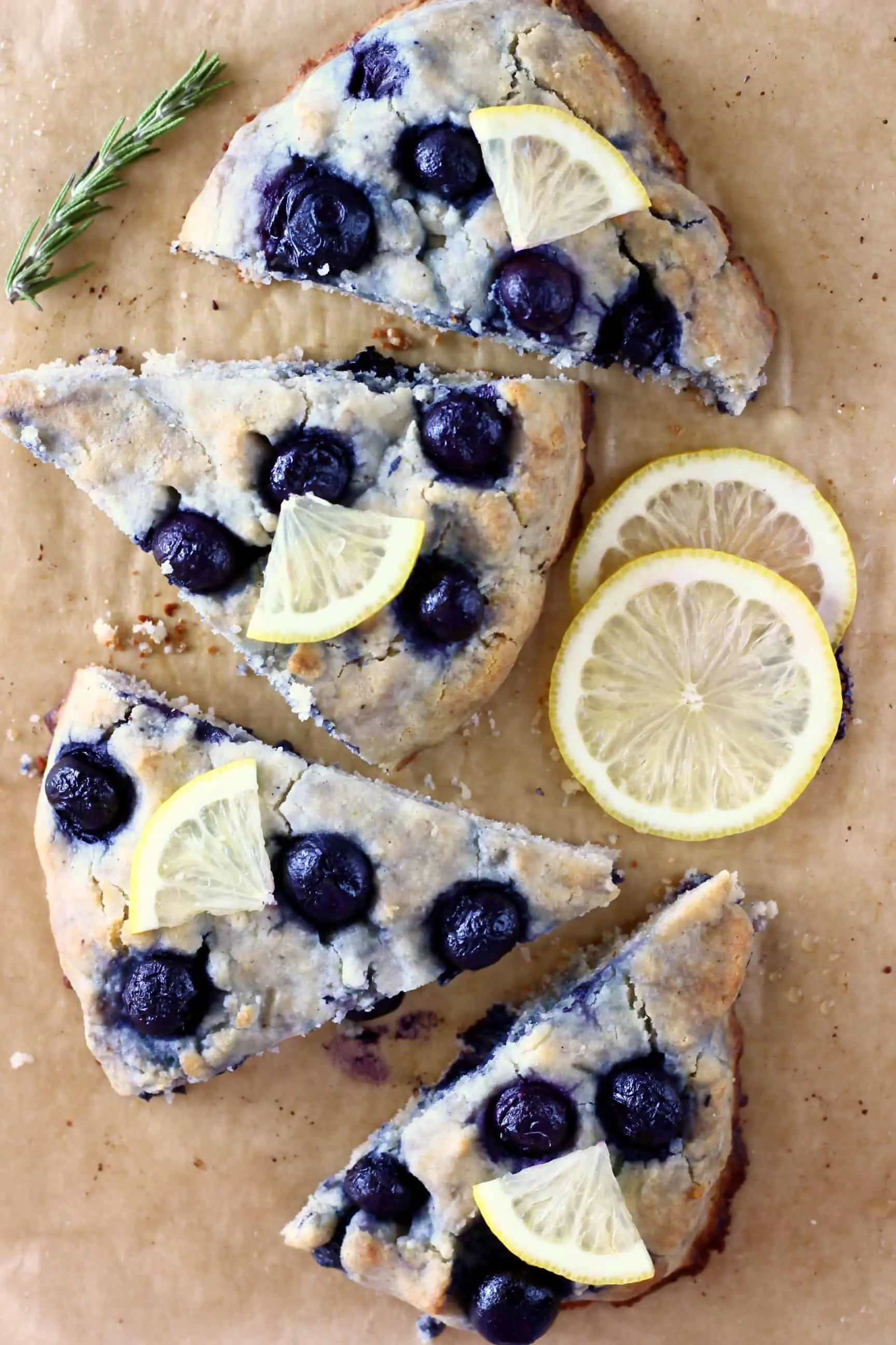Four triangular gluten-free blueberry scones topped with lemon wedges on a sheet of brown baking paper