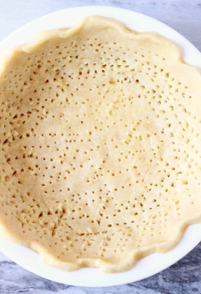 Photo of a raw pastry crust with lots of fork holes in a white pie dish against a marble background