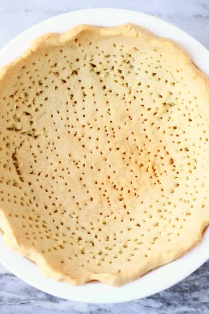 Photo of a cooked pastry crust with lots of fork holes in a white pie dish against a marble background