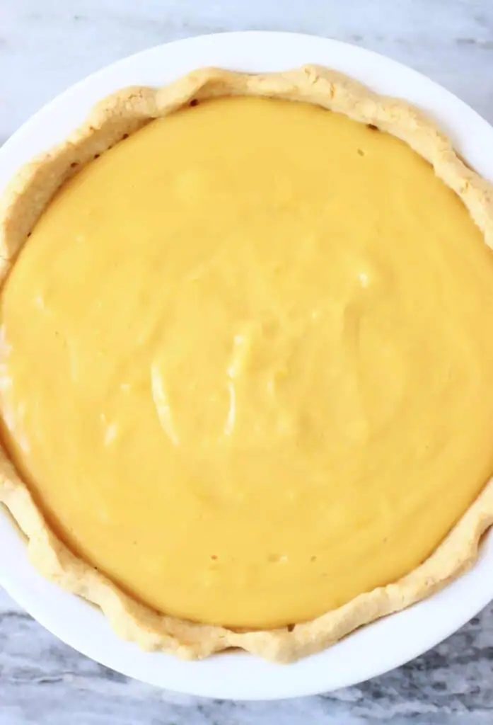 Photo of a pastry crust filled with yellow custard in a white pie dish against a marble background
