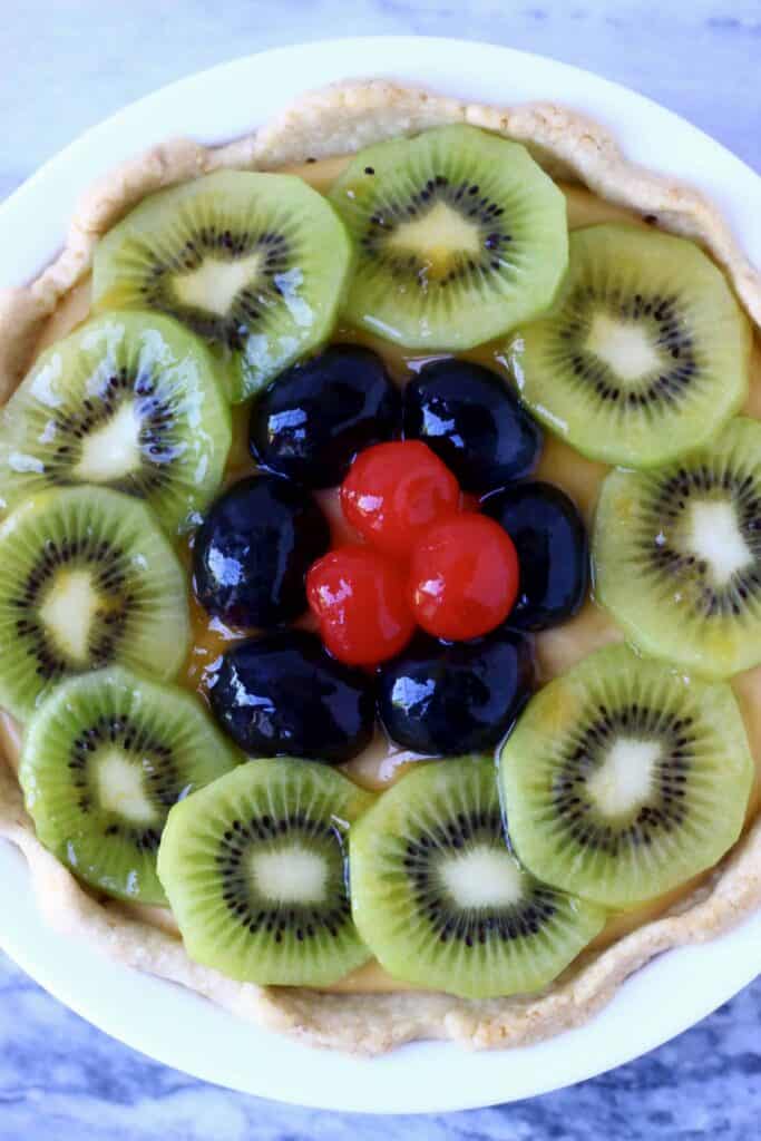 Photo of a fruit tart topped with sliced kiwis, grapes and red cherries in a white pie dish