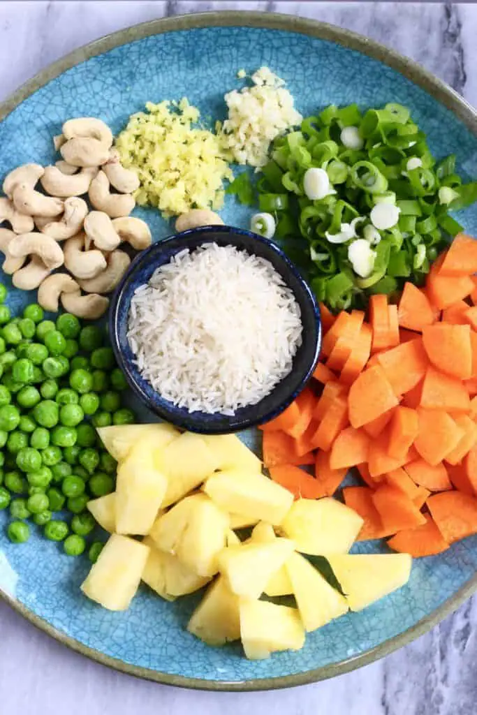 Photo of minced ginger, garlic, sliced spring onions, cashew nuts, green peas, diced carrots, pineapple chunks and raw rice on a blue plate against a marble background